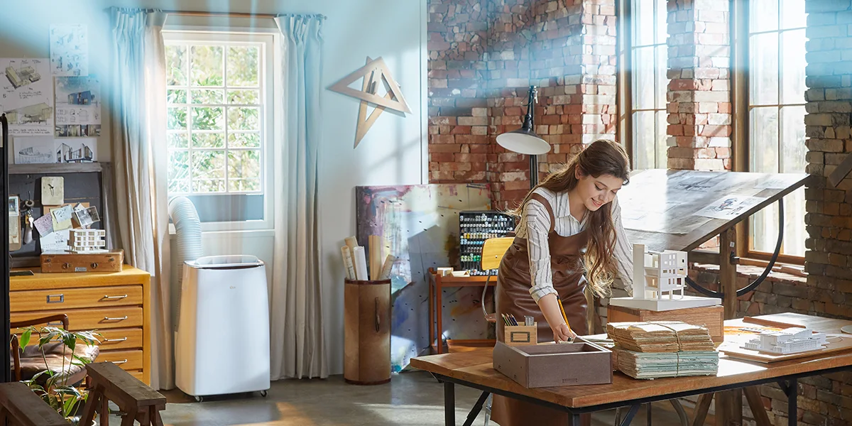 A woman building something in her studio while an air conditioner behind her pumps cool air into her home