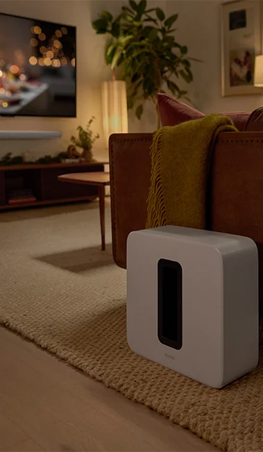Close up on the Sonos Sub4 Subwoofer next to a couch with a dim lit living room ambiance and a tv and sonos soundbar in the far distance of the shot