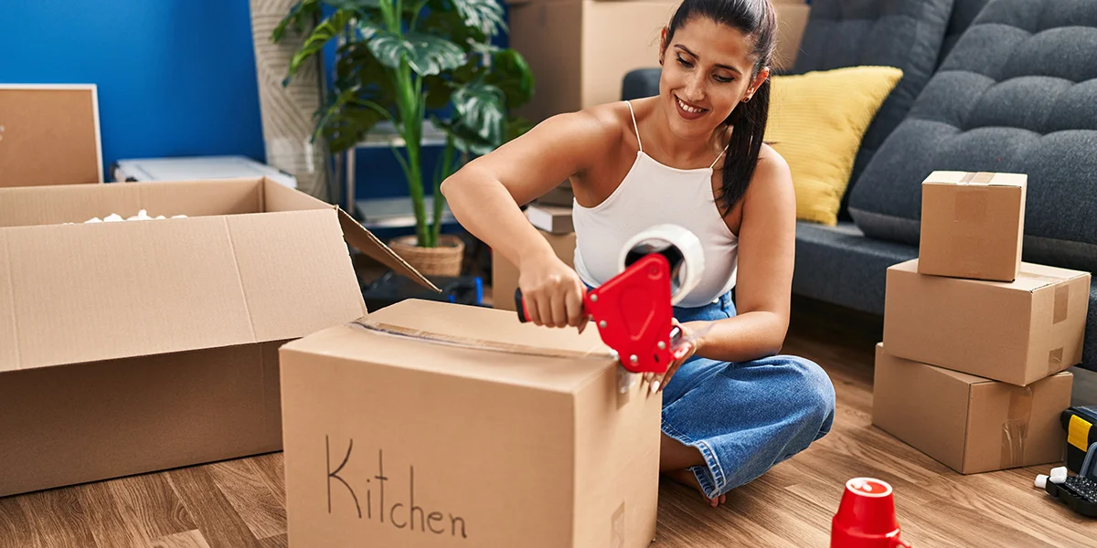 A woman taping a cardboard box shut. The cardboard box is labeled with the word Kitchen.