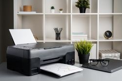 Black Printer situated on a desk in an office setting with a laptop to its right and a bookshelf in the background
