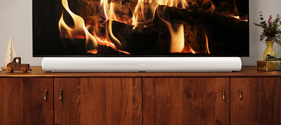 A white Sonos Arc soundbar sitting on a wooden TV stand underneath a flat screen TV that's displaying an image of wood logs burning