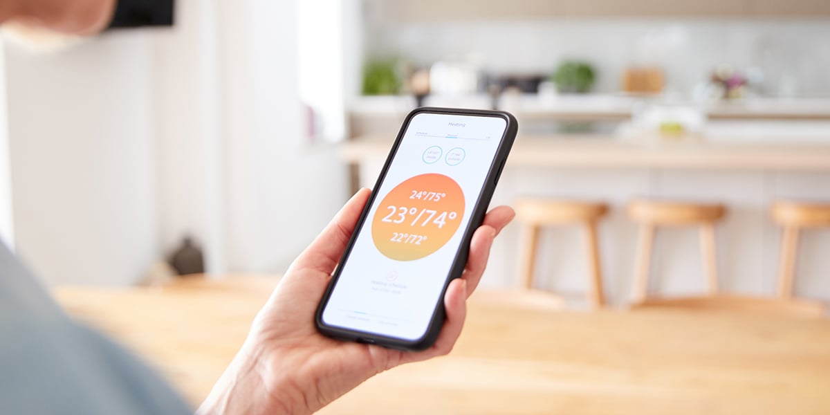 A person holding their smartphone, which shows a smart thermostat app with the temperature at 74 degrees.