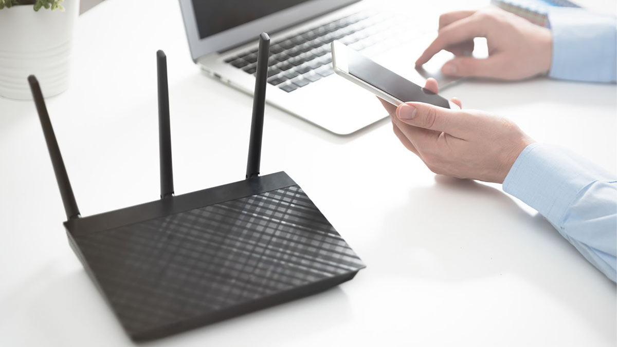 A black router sits on a white table. A person sits at the table, holding a white smartphone in their left hand and controlling a laptop mouse with their right hand