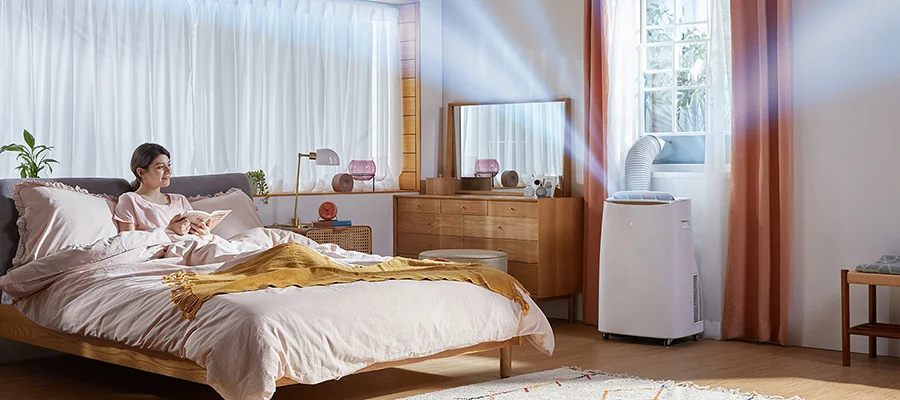 A woman reading in bed while a portable air conditioner releases cool air from the window in her bedroom