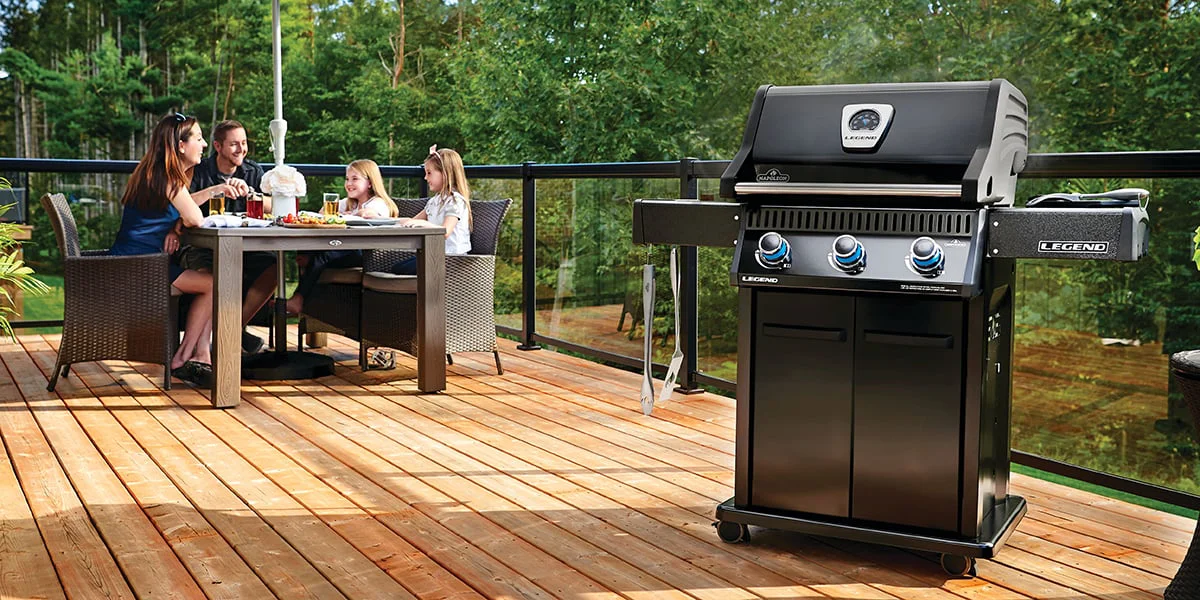 A black outdoor grill standing on a wooden porch while a family eats at a table behind it