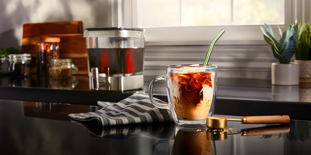 A clear glass mug of coffee with cream sitting on a counter while a coffee maker sits in the background