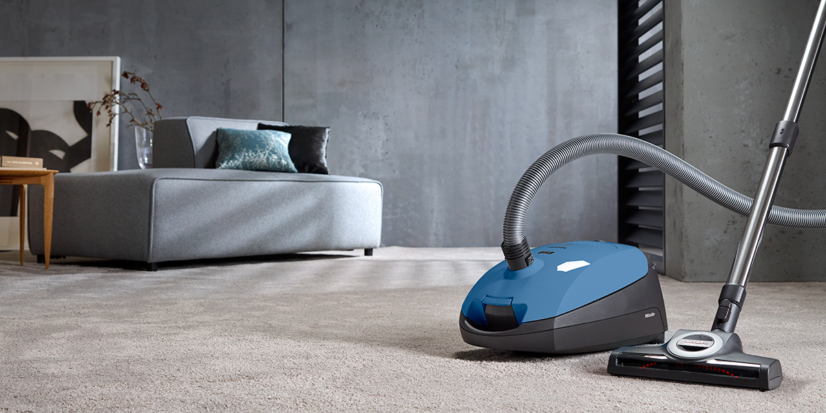 A blue vacuum cleaner sitting on a living room carpet with a sofa and table behind it