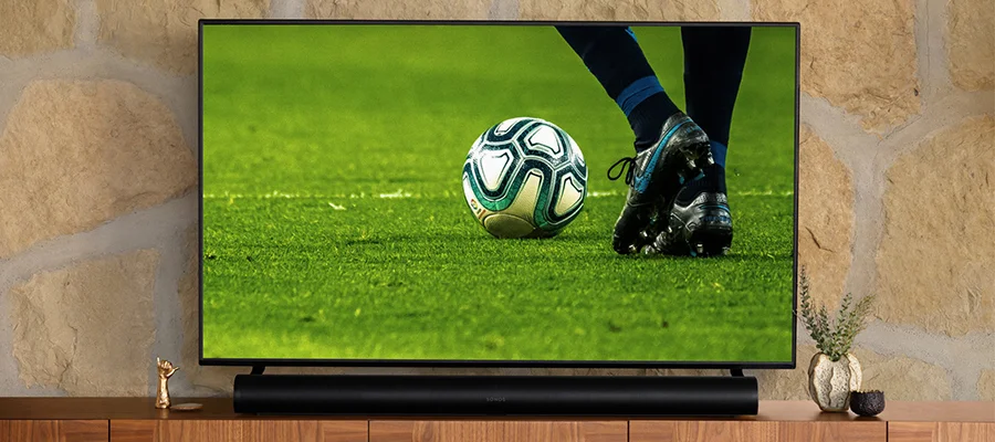 A black Sonos Arc soundbar resting on a wooden TV stand underneath a flat screen TV that's displaying a soccer game