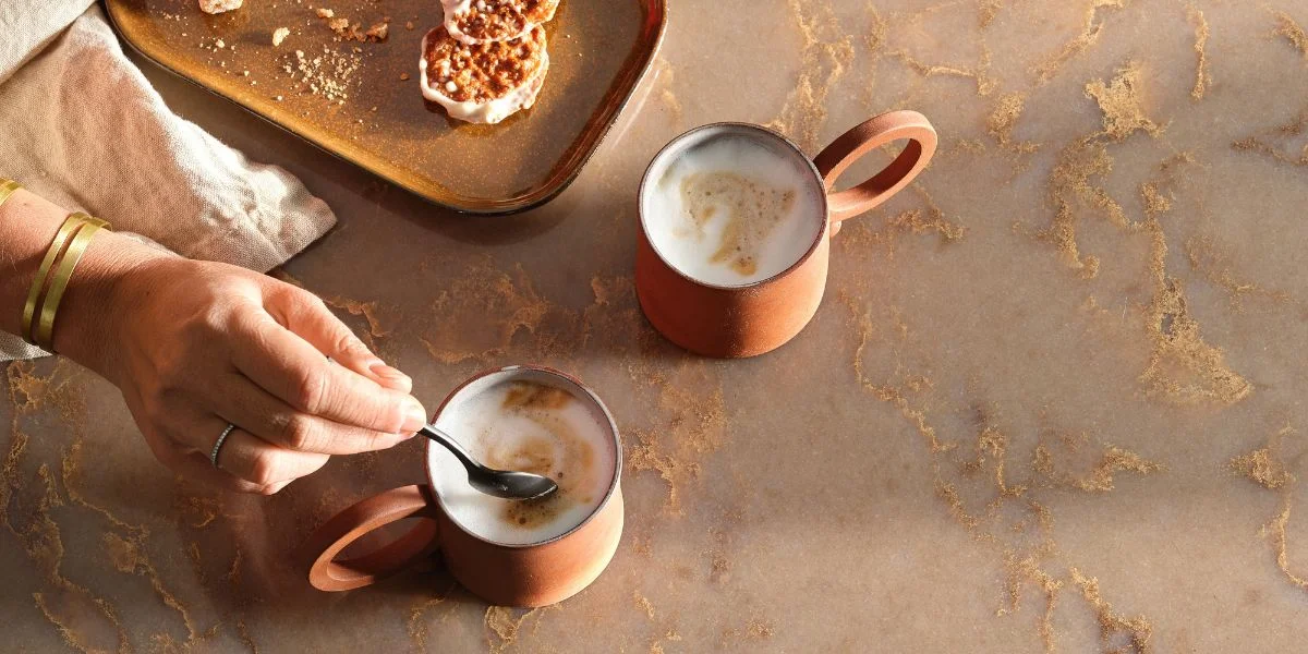 Two cups of coffee topped with frothed milk beside some miniature pancakes. A woman is stirring her beverage with a spoon.
