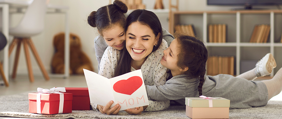 A mom reading a mother's day gift while her two daughters snuggle her on the floor