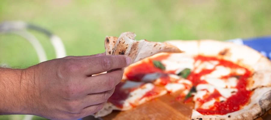 A close-up of a hand holding a slice of pizza outdoors.
