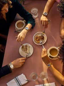 over head shot of a people at a table with cake and coffee and varoups watches on the wrists of the people at the table
