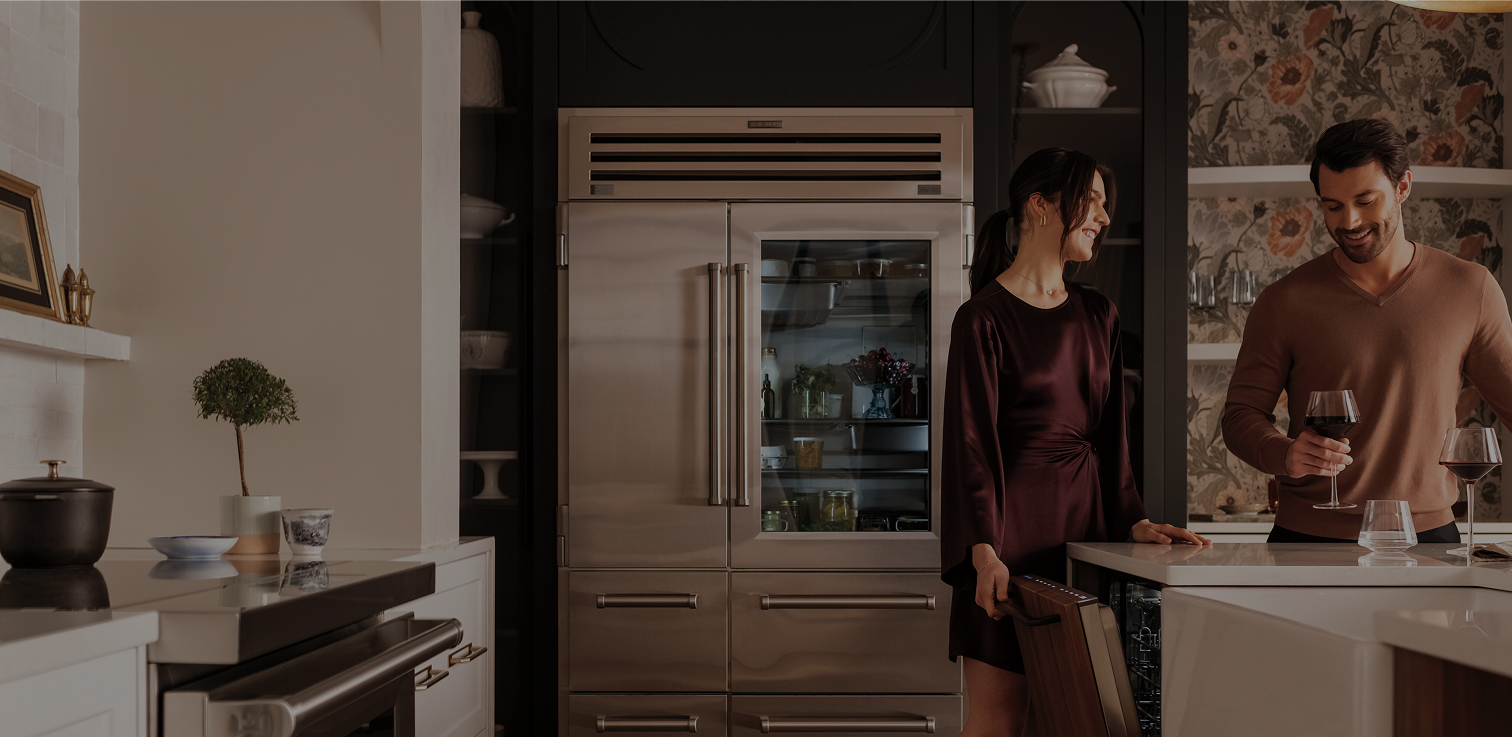 A man and woman stand together in a kitchen, next to a sub-zero refrigerator and dishwasher engaged in conversation. She is opening the dishwasher