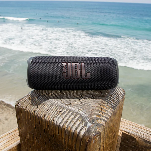 A black JBL Flip 6 speaker sitting on a wooden post by the beach