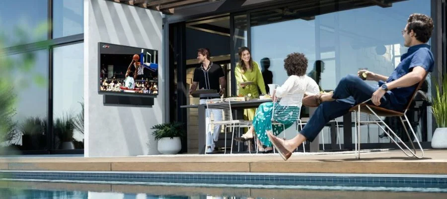 An image of friends gathered around a poolside table watching an outdoor TV and enjoying lunch.