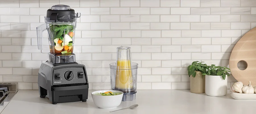 A black Vitamix blender filled with fruits and vegetables sitting on a white kitchen counter next to a bowl and a personal blender jug