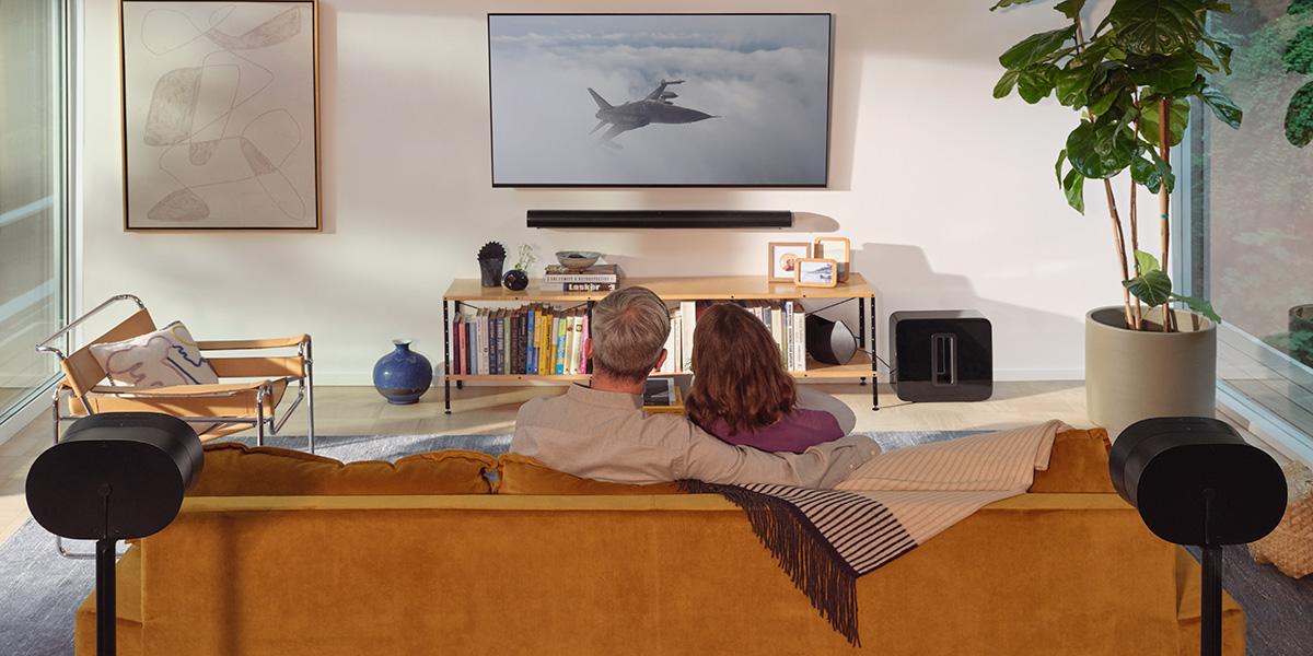 A pair of people watching television with two Sonos speakers behind them providing Dolby Atmos sound