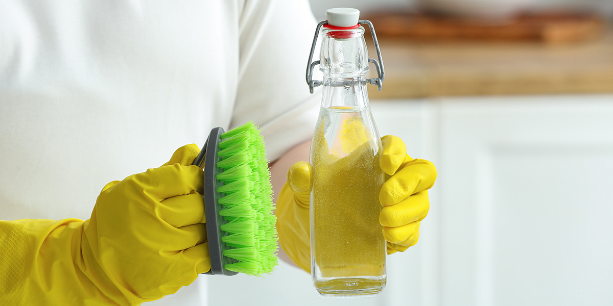 A person wearing yellow cleaning gloves holding a scrubbing brush and a bottle of white vinegar