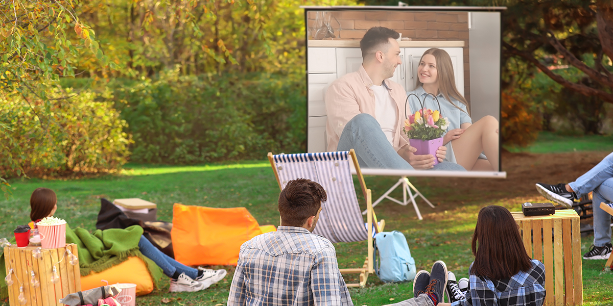 People sitting outside on the grass or in chairs watching a projector screen with an image of a couple