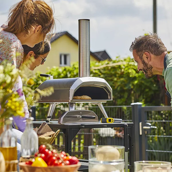 A pizza oven sitting on a table outside while people look in to see the food