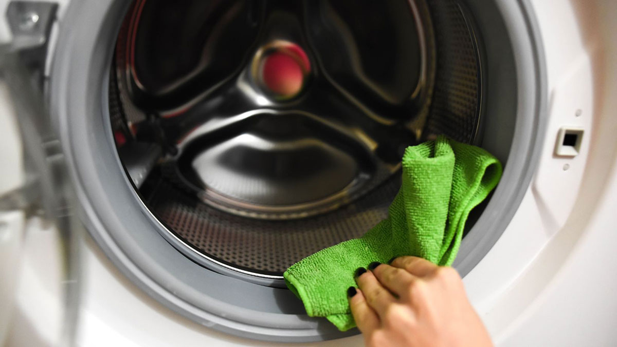 Person's hand holding a green microfiber cloth cleaning the inside of a front load washer
