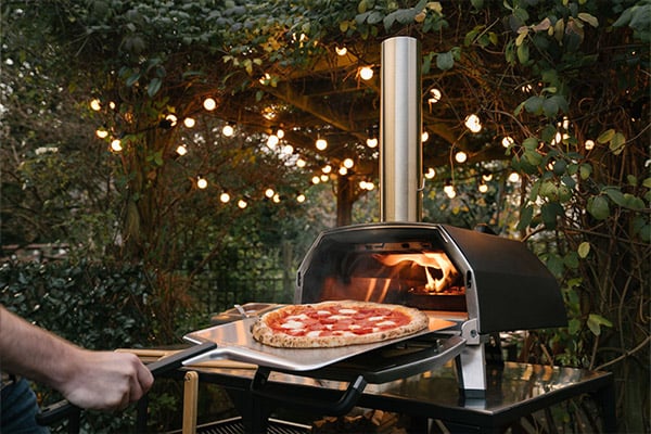 Person removing a cheese pizza from a tabletop Ooni oven at night