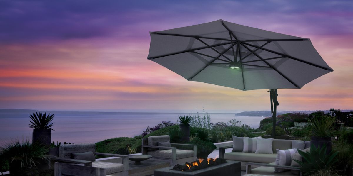 A tilted umbrella over an outdoor seating area. A light is attached and there's a fire beside the chairs and couches