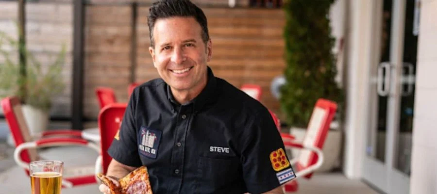 "The Food Guy" Steve Dolinsky sitting at a table with a pizza in front of him. He is looking at the camera and smiling