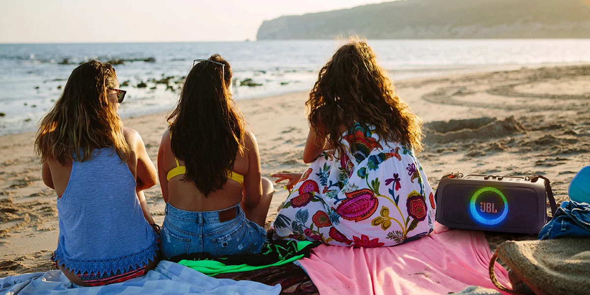 Three girls sitting on a beach beside a JBL partybox speaker in the sand
