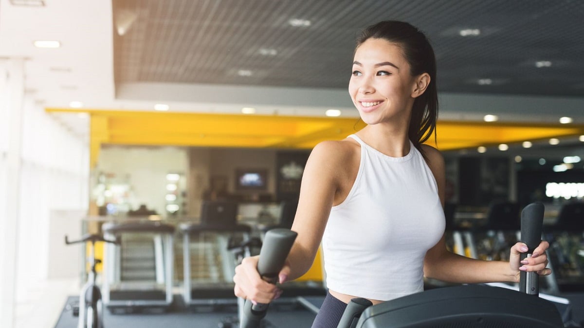 Woman with brown hair wearing a white racerback top on an elliptical holding the hand grips