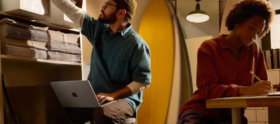 A man on a stool using a macbook air with one hand. A woman sits at a table behind him