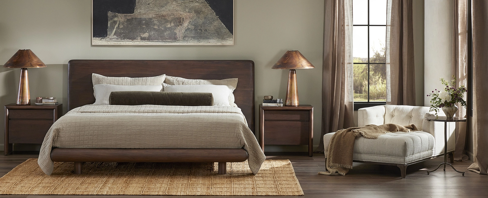 Warm, contemporary bedroom interior with a large upholstered bed dressed in neutral bedding, two matching wood nightstands with lamps, a leafy potted plant near the window, and two brown lounge chairs in the corner.