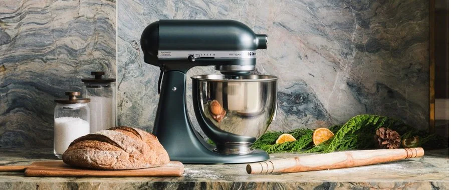 The KitchenAid Artisan Plus stand mixer sitting next to a fresh loaf of bread and rolling pill, all on a festive countertop.