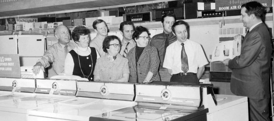 A group of Abt salespeople in a black and white photo surrounded by appliances as they learn about the latest products to sell.
