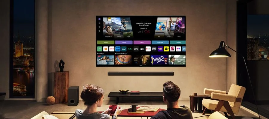 An LG G5 wall-mounted TV in a living room with two people watching. The room is dim and it is dark outside. The two are choosing what to watch on the G5 smart TV