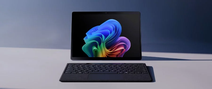 A 13-inch Microsoft Surface Pro 2-in-1 PC on a table with a detached wireless keyboard. The screen shows a Windows 11 background, a folded floral display.