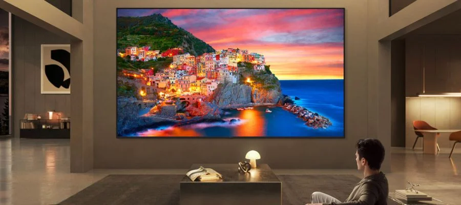 A man watching the TV at home in a gray living room. The TV is displaying a sunset on an island with a beautifully-lit town.