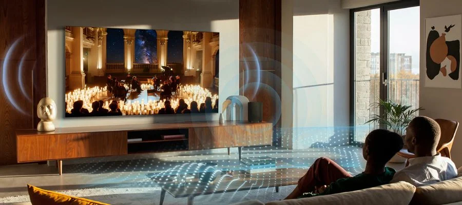 A couple on a sofa watching a candlelit orchestral concert onscreen. Waves of sound are coming from the TCL TV and surround the viewers.