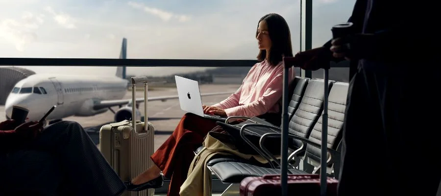 A woman using a new MacBook Pro with an M5 chip in an airport. An airplane is outside the window.