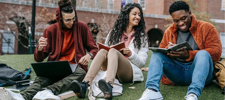 Three students sitting outside and studying together with notebooks and computers.
