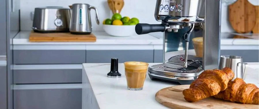 The Breville Bambino Plus sitting on a modern kitchen island next to a fresh espresso shot and croissant.