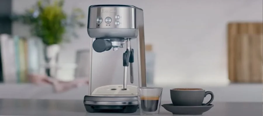 The Breville Bambino sitting on a counter next to a fresh shot of espresso and a mug of coffee.
