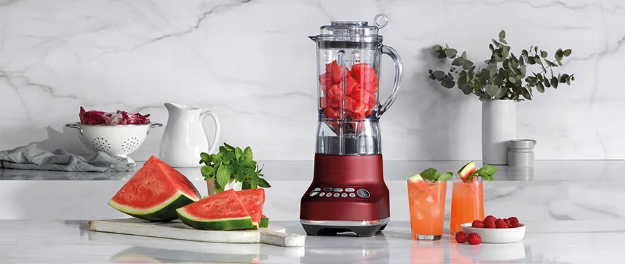 A red Breville blender filled with watermelon chunks sitting on a kitchen counter next to slices of watermelon, raspberries and two iced beverages