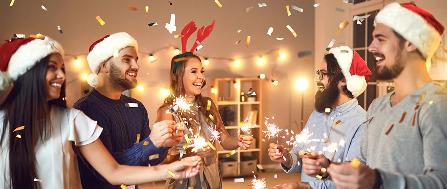 A group of friends wearing Santa hats and holding sparklers as silver and gold confetti falls around them