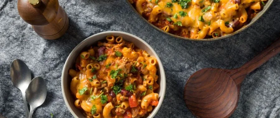 A bowl of chili mac and a serving platter off to the side, along with two spoons, a pepper mill and a wood serving spoon