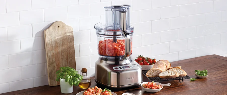 The Breville the Paradice 16 food processor sitting on a wooden countertop surrounded by ingredients while chopped tomatoes sit in the bowl