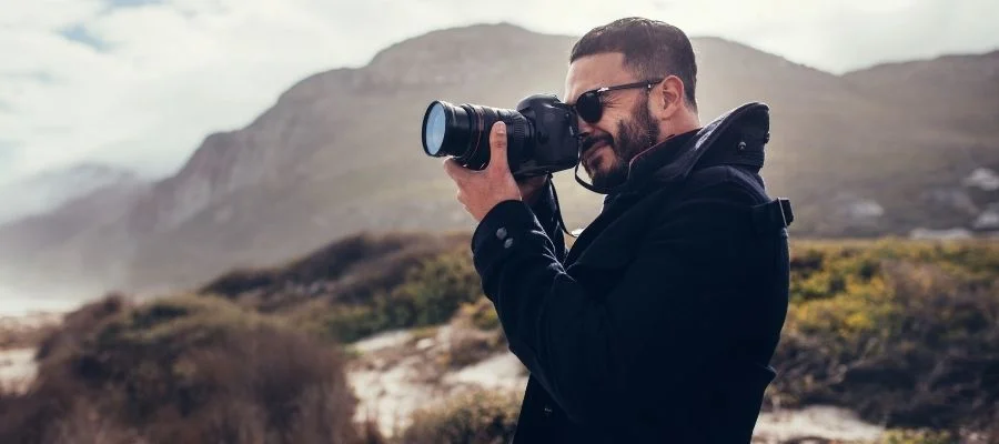 Man wearing sunglasses taking a photograph on grasslands with a mountain in the background.