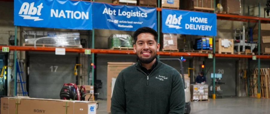 Enrique Hernandez standing in the warehouse at Abt in front of Logistics banners that read 'Abt Nation' 'Abt Logistics' and 'Abt Home Delivery'