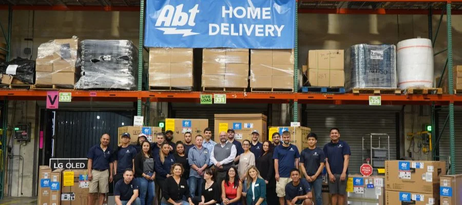 A zoomed-out photo of the Abt logistics team in the warehouse beneath a 'Home Delivery' sign