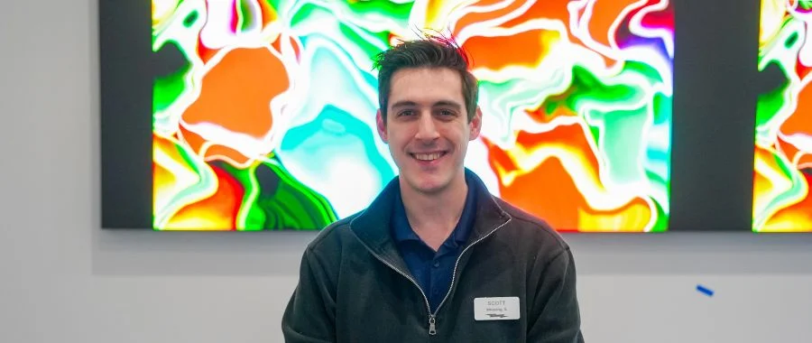 Scott Grigoletto smiling in front of a colorful television in a meeting room.